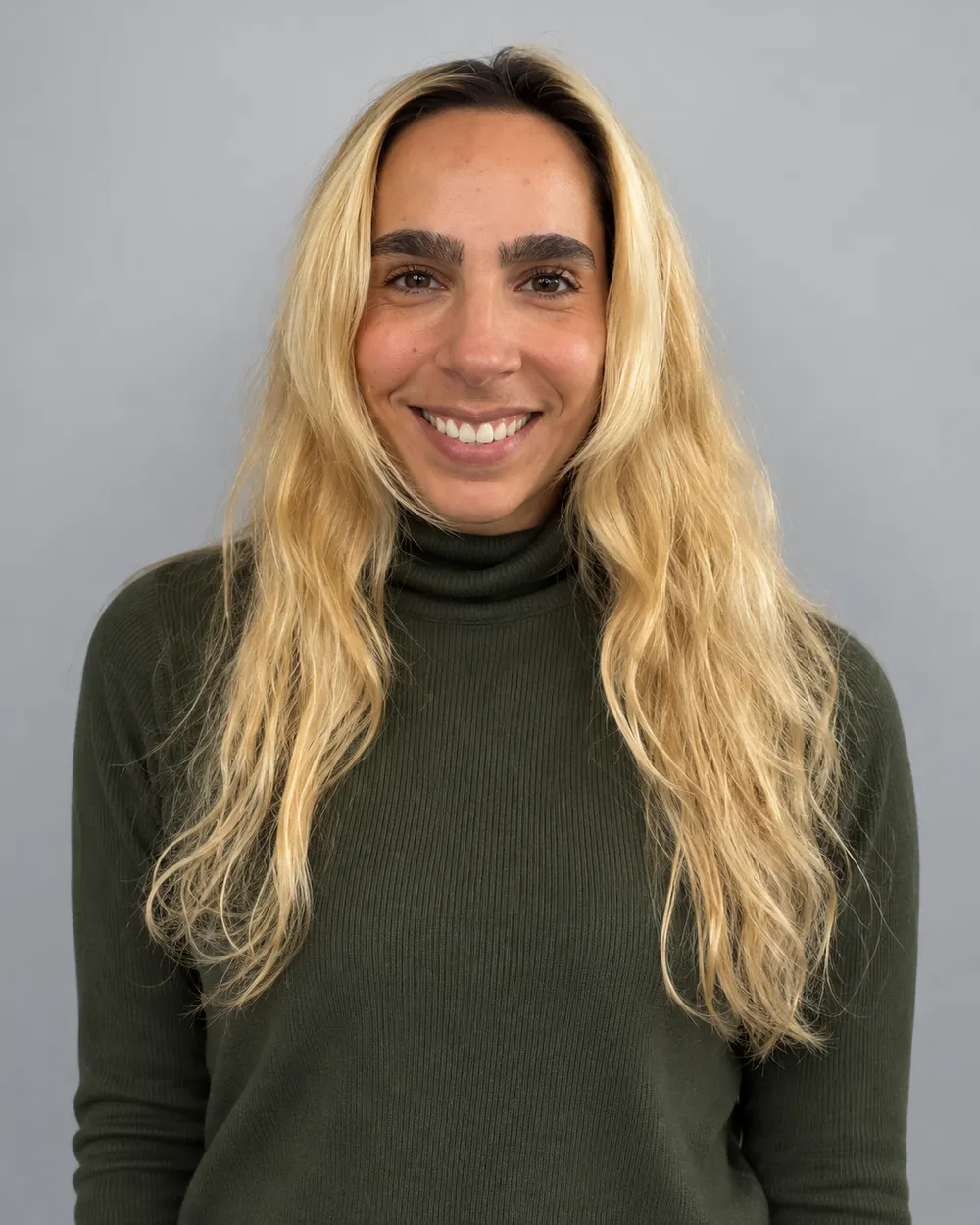 Mulher sorrindo com cabelo longo ondulado loiro e raiz natural, usando blusa verde de gola alta sobre fundo cinza