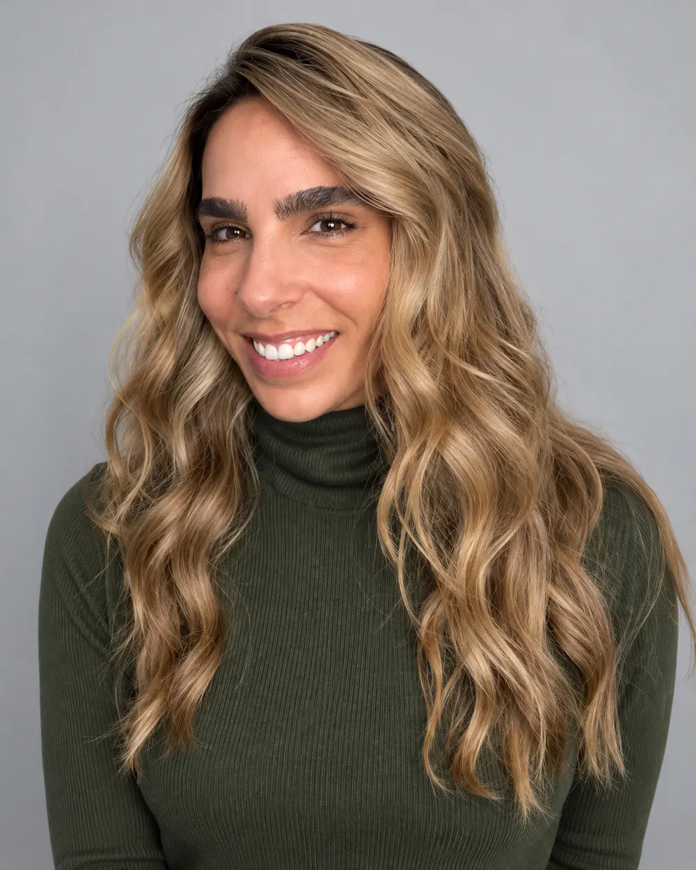 Mulher sorrindo com cabelo longo ondulado loiro e raiz natural, usando blusa verde de gola alta sobre fundo cinza